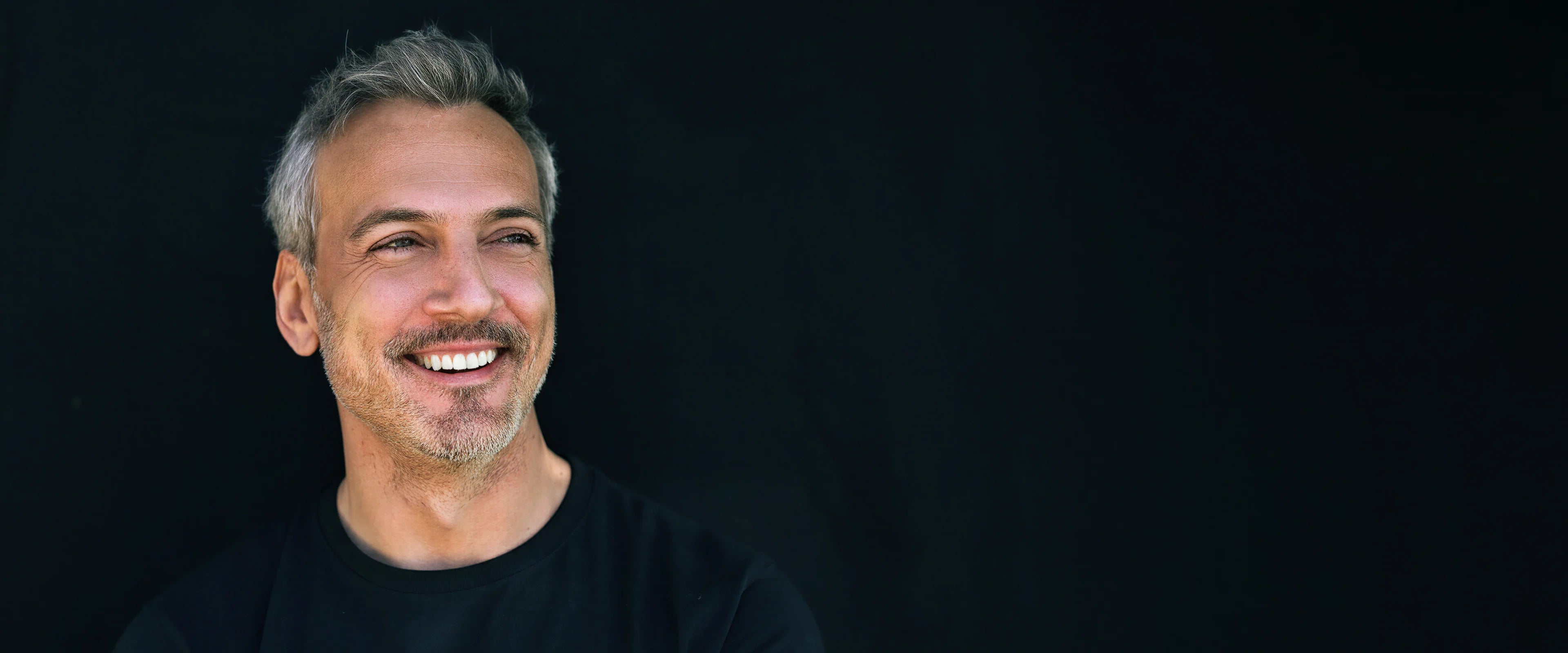 Lächelnder Mann mit grauem Haar vor dunklem Hintergrund, natürliches Motiv für Männerhautpflege