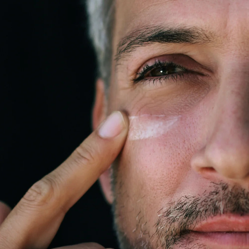 Nahaufnahme eines Mannes, der eine Augenpflege oder Creme sanft unter sein rechtes Auge aufträgt; sein Finger verteilt die weisse Textur auf der Haut, während sein Blick leicht seitlich gerichtet ist.