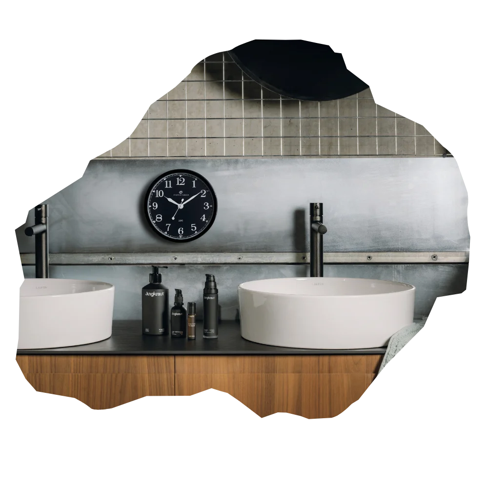 Modernes Badezimmer mit zwei runden, weißen Waschbecken auf einer dunklen Ablage; dazwischen stehen mehrere schwarze Jungkraut-Pflegeprodukte. Im Hintergrund sind Metallfliesen und eine schwarze Wanduhr sichtbar.